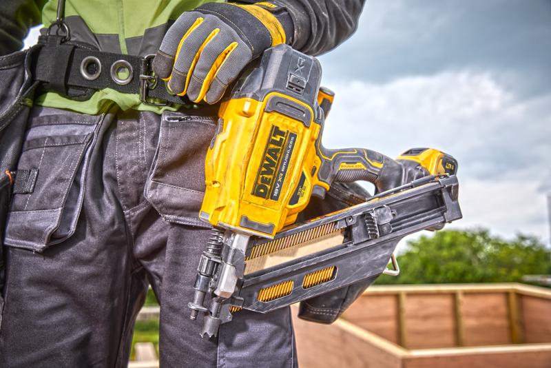 Person holding a DEWALT DCN930N nail gun, wearing work gloves and gear at a construction site outdoors.